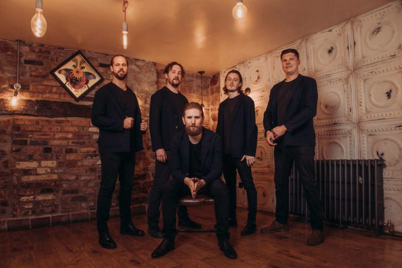 All male function band stood in quirky dim lit room all dressed in black suits