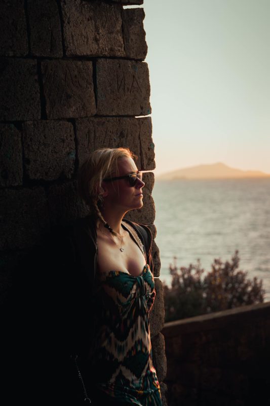 A girl leaning against a brick wall stood staring into the sun setting over the Gulf of Naples.