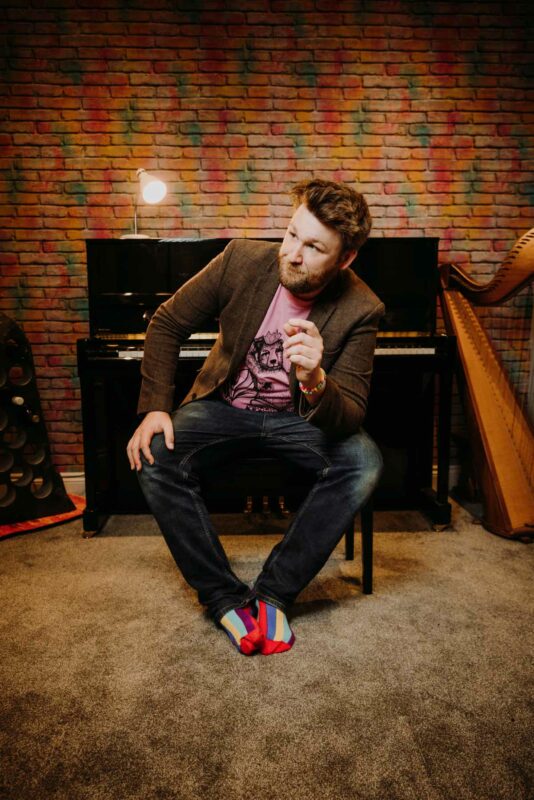 Portrait of Greg Ryan, musician, sat in front of a piano. Shot as part of a recent promo shoot at his home studio in Newport.