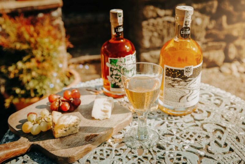 A glass of Hive Mind traditional mead sat on a table in a sunny garden, surrounded by cheese.