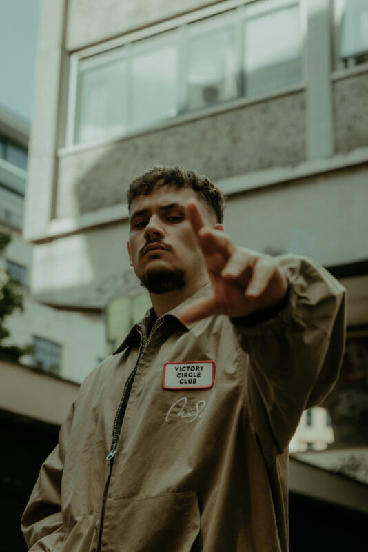 Hip hop artist and rapper SAINT stood in an urban area of Bristol, gesturing to the camera with a high rise building behind.