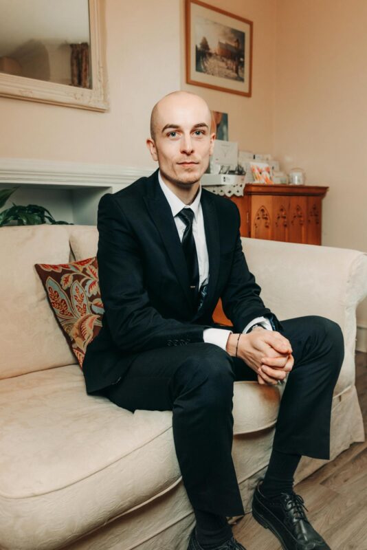 Portrait of a funeral director sat in a black suit smiling solemnly on a cream sofa. Shot for a marketing rebrand in October 2024.