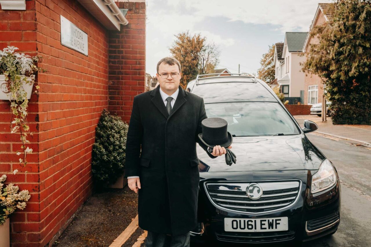 Portrait of a funeral director stood next to a hearse holding a funeral hat. Shot for a marketing rebrand in October 2024.