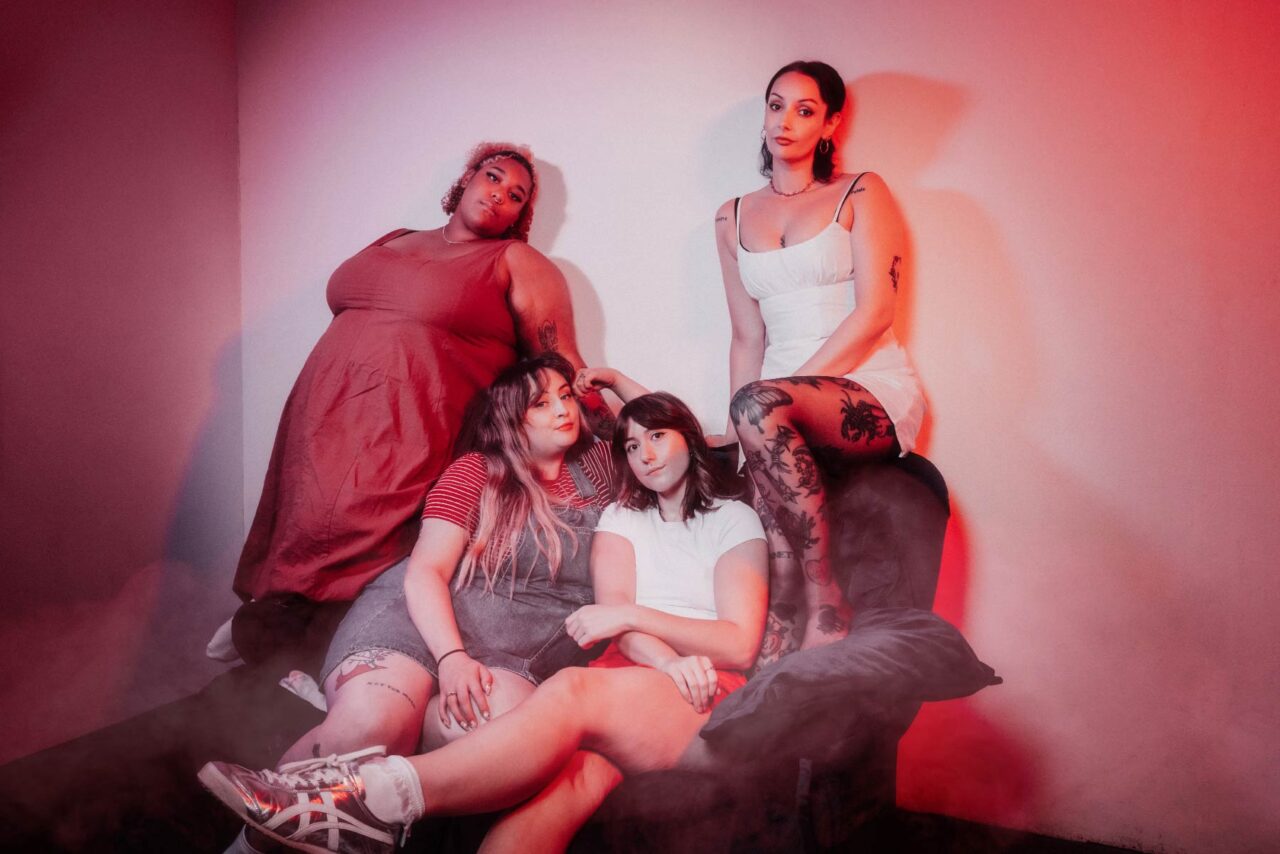 Pop rock girl band Murder Club sat on a sofa surrounded by red light and smoke. Shot at One Louder Studios, Newport.