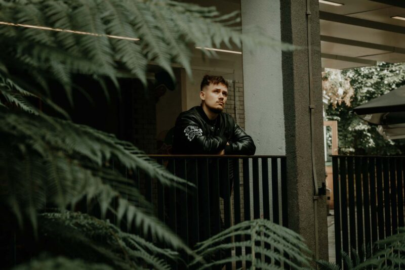 Urban rapper leaning against some railings, framed by some fern leaves. Shot in Bristol city, summer 2024.